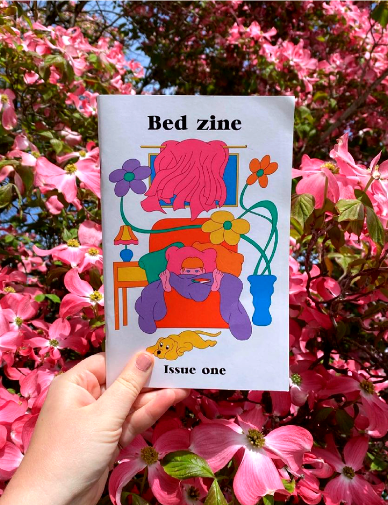 This is an image of a hand holding up a copy of Bed Zine's front cover. Featuring a brightly coloured illustration of a person lying in bed. Behind the hand is a beautiful backdrop of bright pink flowers.