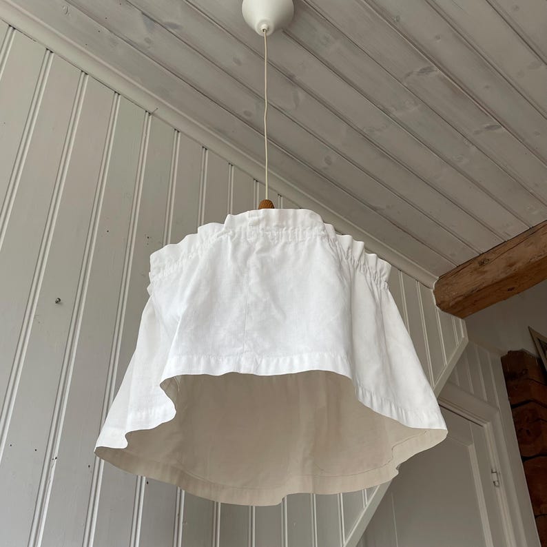 May include: A white fabric lampshade with a ruffled design, hanging from a white ceiling fixture. The lampshade has a unique, layered shape and is suspended by a thin cord. May include: A white fabric lampshade with a ruffled design, hanging from a white ceiling fixture. The lampshade has a unique, layered shape and is suspended by a thin cord.