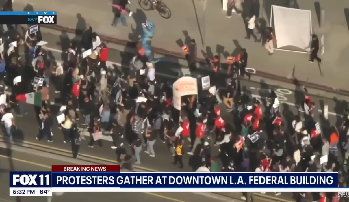 Video screenshot, helicopter view of a protest march outside a Los Angeles federal building where immigrants held in ICE raids were detained. Marchers carry signs and banners, and at the top center of the image is one marcher wearing an inflatable blue T Rex dinosaur costume, its head reared as if roaring (but probably to allow the wearer to see where they're going) Video screenshot, helicopter view of a protest march outside a Los Angeles federal building where immigrants held in ICE raids were detained. Marchers carry signs and banners, and at the top center of the image is one marcher wearing an inflatable blue T Rex dinosaur costume, its head reared as if roaring (but probably to allow the wearer to see where they're going)