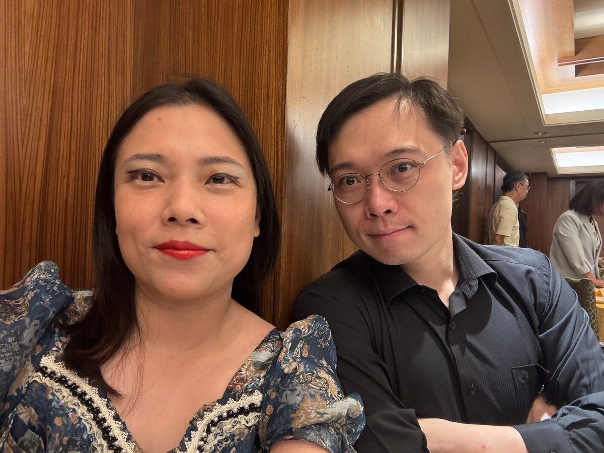 Huang Shih-shiow and a woman with long dark hair wearing a blue floral dress with lace trim sit side by side against a wooden panel wall. Huang Shih-shiow, wearing glasses and a dark shirt, has his arms crossed. The background shows a softly lit indoor setting with other people visible in the distance.
