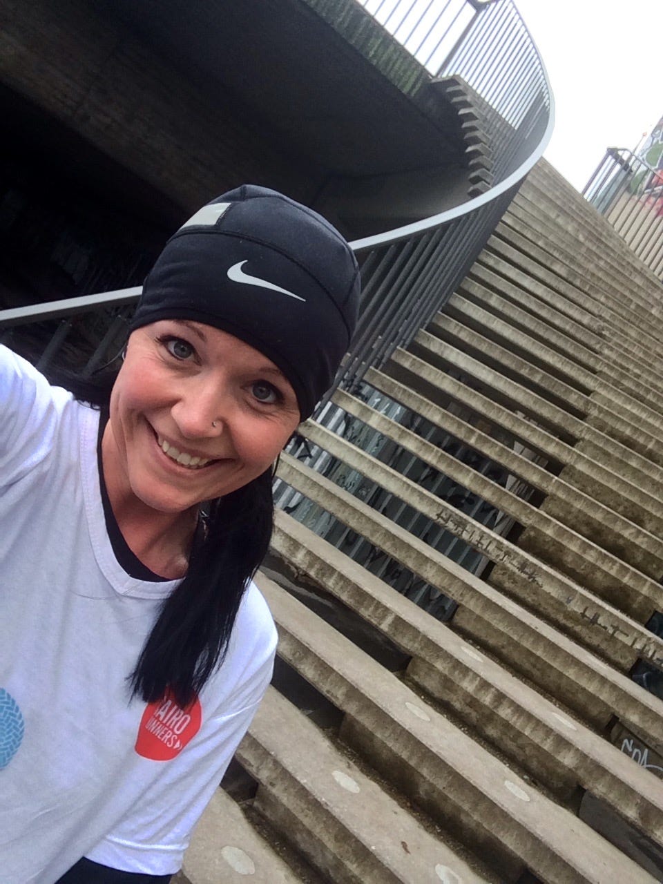 Georgina Dean, wearing a running beanie, takes a smiling selfie on a spiral concrete staircase in Berlin, Germany, representing a reckless and joyful creative journey.