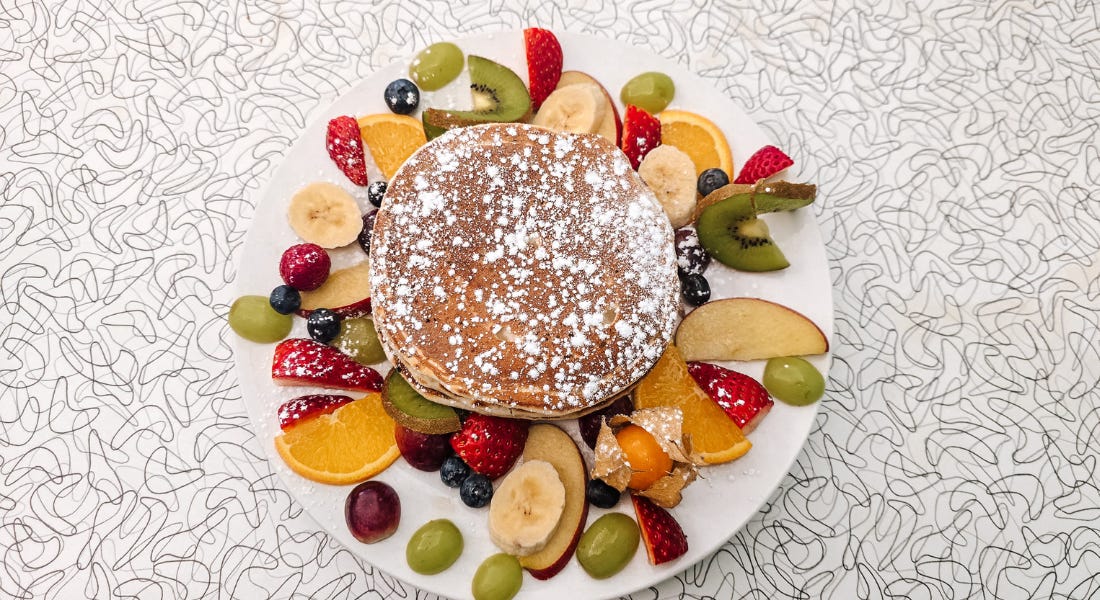 A plate of pancakes dusted with icing sugar with fruit around them, on top of a black and white abstract surface