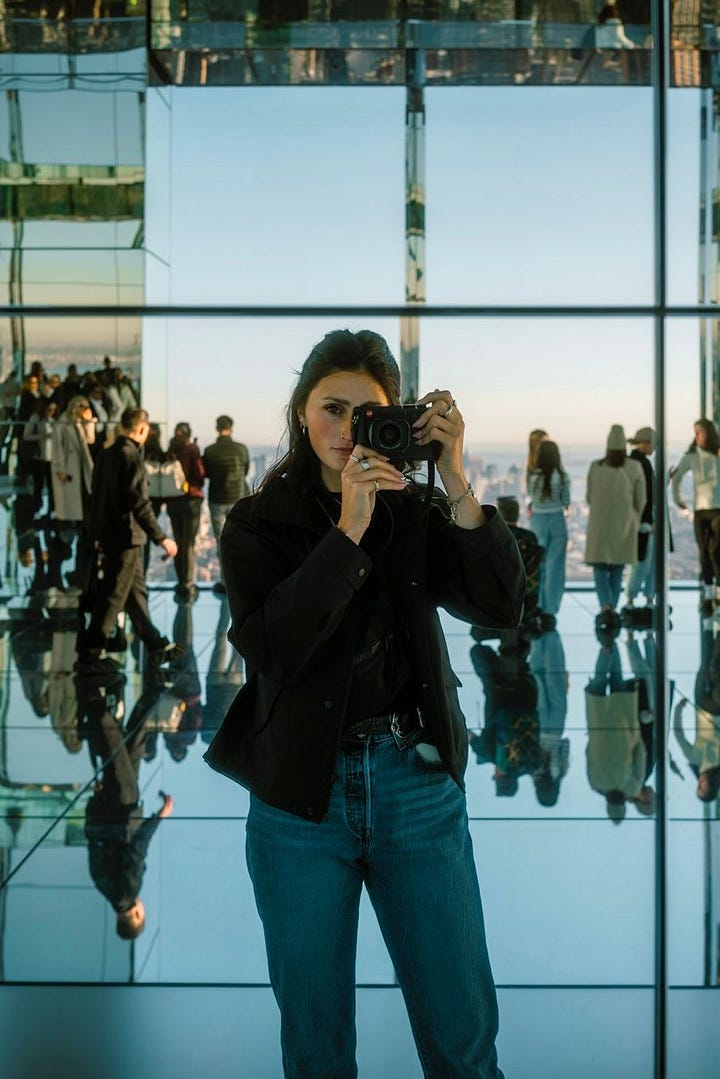 Two photos of a woman with a camera: one is a smiling selfie taken on a rocky beach with cliffs behind her, and the other shows her photographing herself in a mirrored indoor space with people reflected around her.