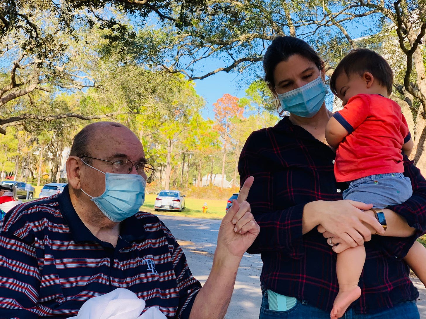 An old man wearing a surgical mask points at a shy baby, held by his mom, whose eyes are closed because it's sunny.