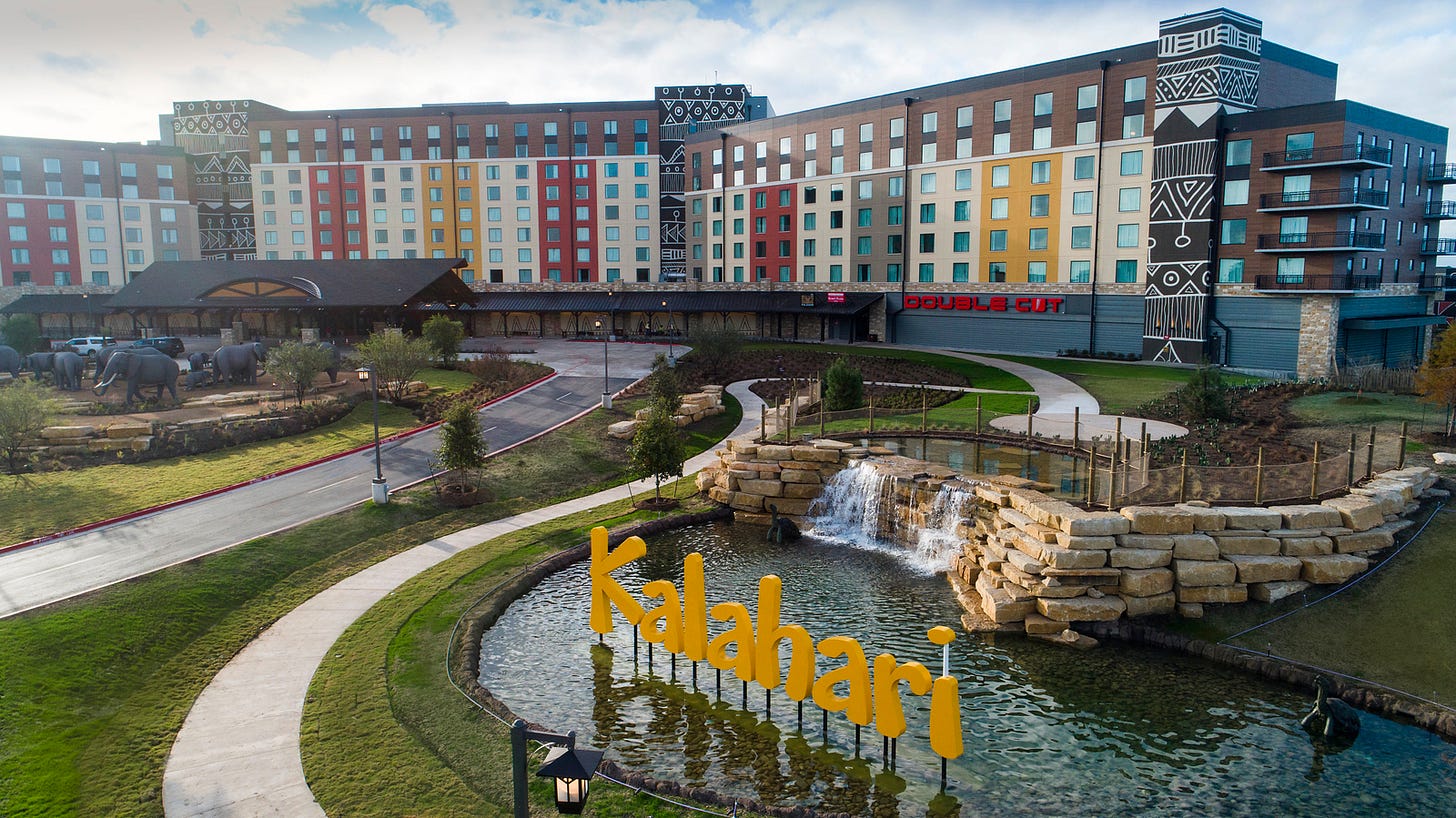 Exterior view of Kalahari Resort in Round Rock, Texas, featuring a colorful multi-story hotel building with African-themed designs, a front drive with elephant statues, a landscaped pond with a yellow “Kalahari” sign, and a small waterfall feature.