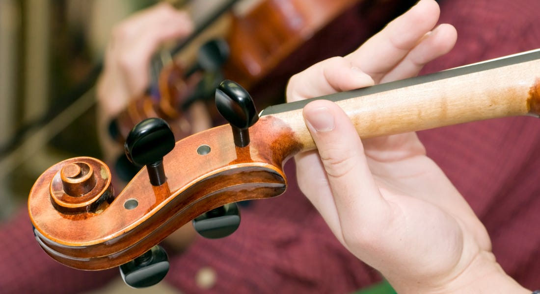 A close-up shot of a fiddle being held by a person wearing a red button-down shirt, with another fiddle player in the distance
