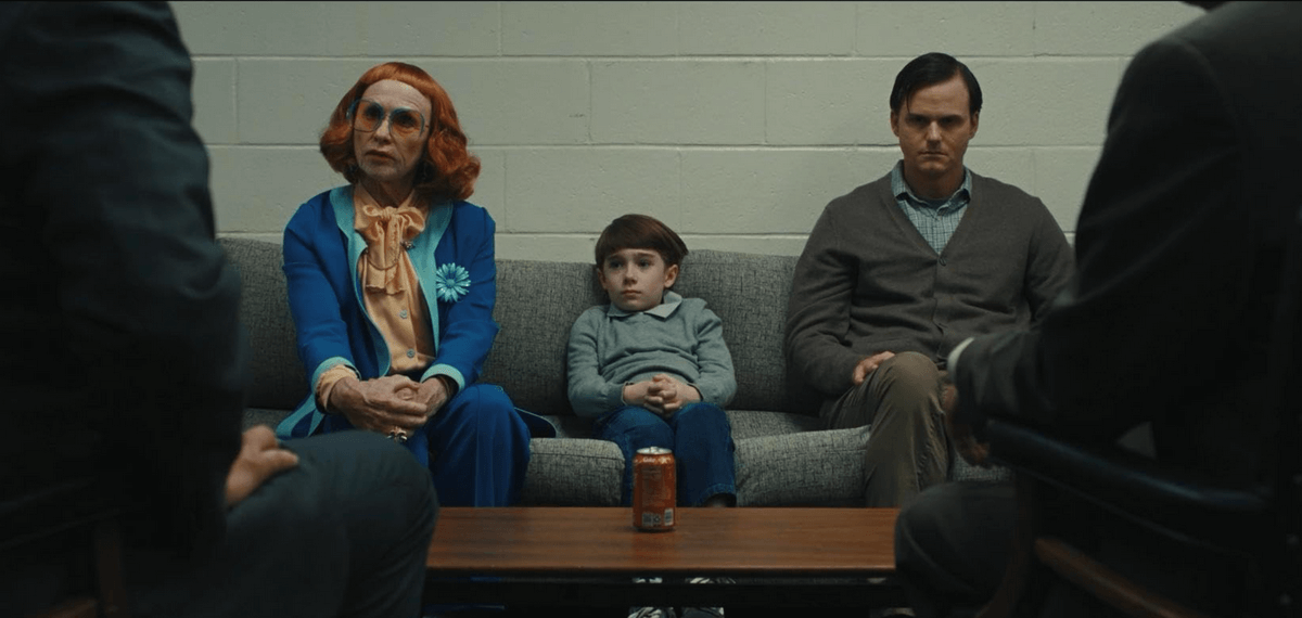 Criança sentada entre dois adultos em uma sala de reunião; à esquerda, mulher de roupa colorida e cabelo ruivo; à direita, homem sério de suéter cinza. Criança sentada entre dois adultos em uma sala de reunião; à esquerda, mulher de roupa colorida e cabelo ruivo; à direita, homem sério de suéter cinza.