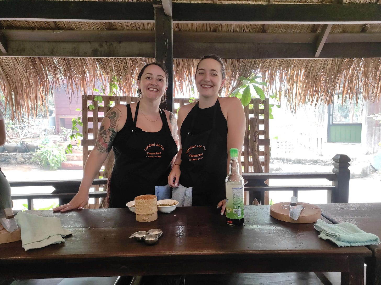 Sophie and Holly smiling at the camera wearing black aprons standing in front of a wooden bench at a cooking class in Laos Sophie and Holly smiling at the camera wearing black aprons standing in front of a wooden bench at a cooking class in Laos