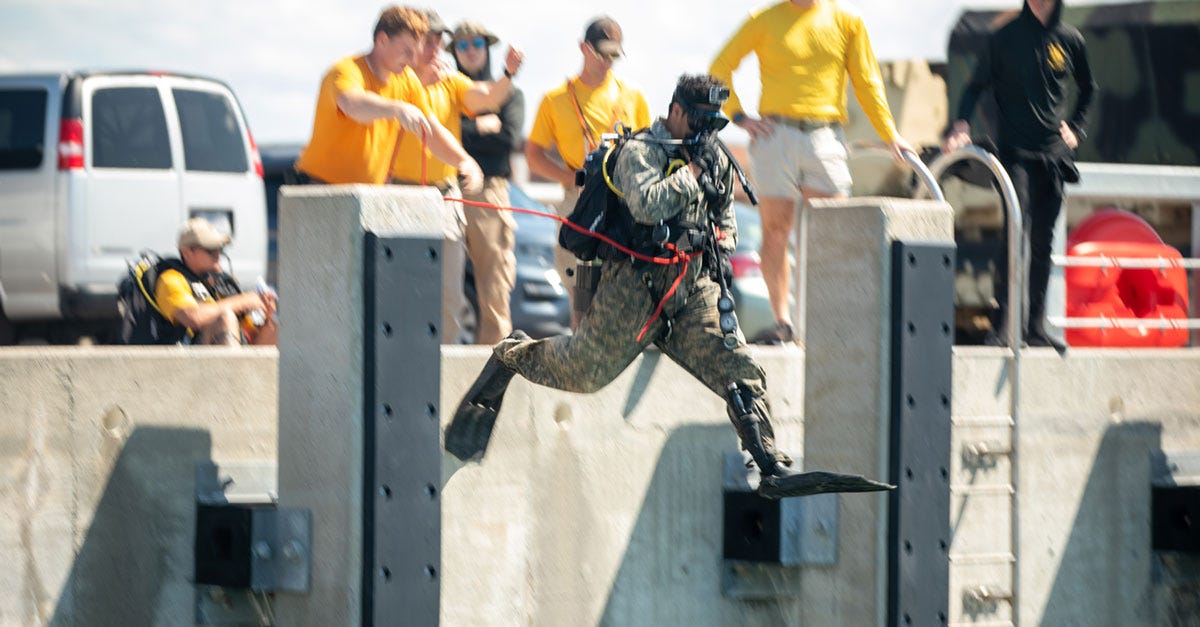 A person in military gear and flippers jumps into the water from a platform, while several people in yellow shirts watch from behind.