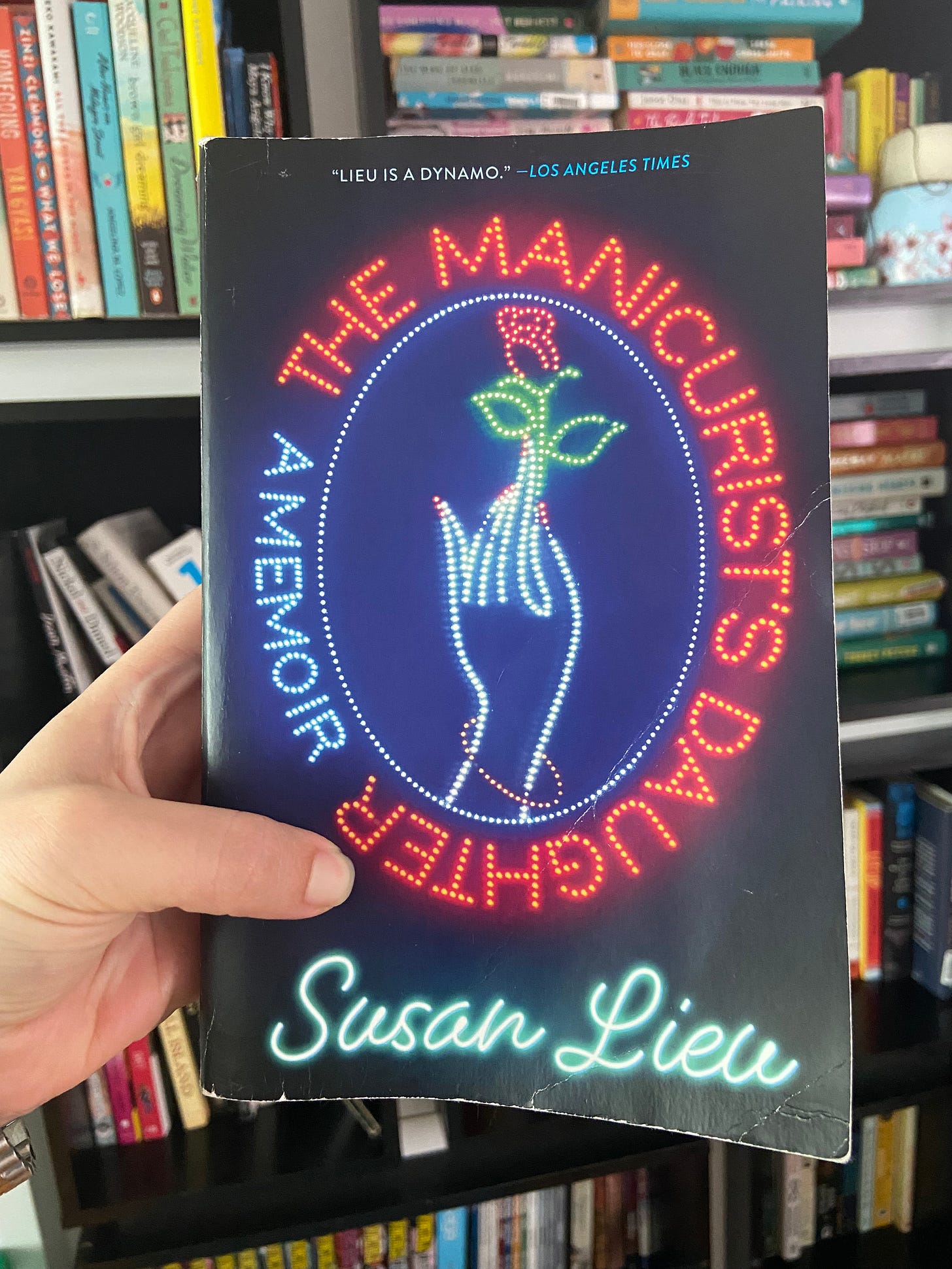 white hand holding up a paperback copy of The Manicurist’s Daughter in front of bookshelves