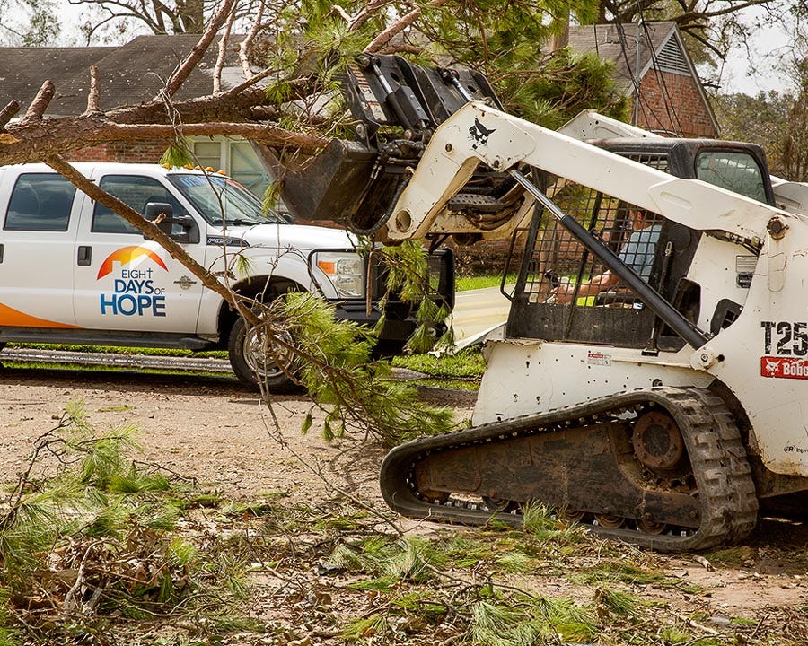 Skidloader moving branches with a white Eight Days of Hope truck parked in the background.