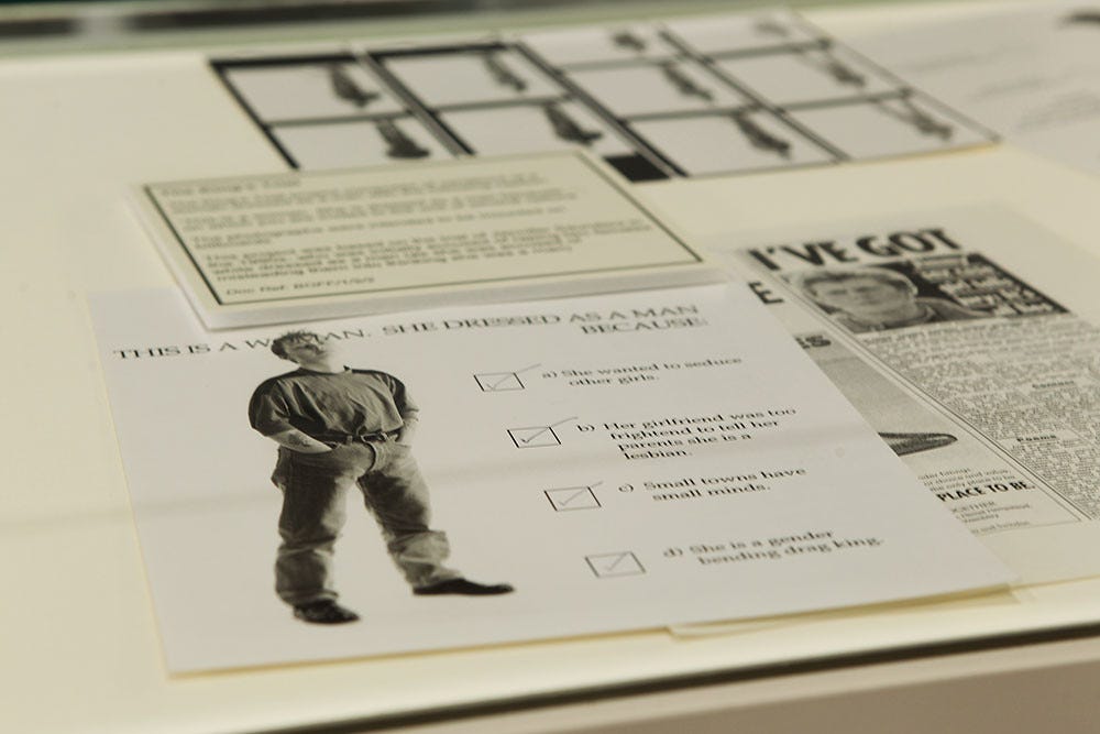 A display case with a paper showing a woman dressed in men's clothing, surrounded by text, checkboxes, and a newspaper. The tone is educational. There is also a text placard behind the image giving context to what all is in the display box. Tessa's inspirations and photographs are displayed together.