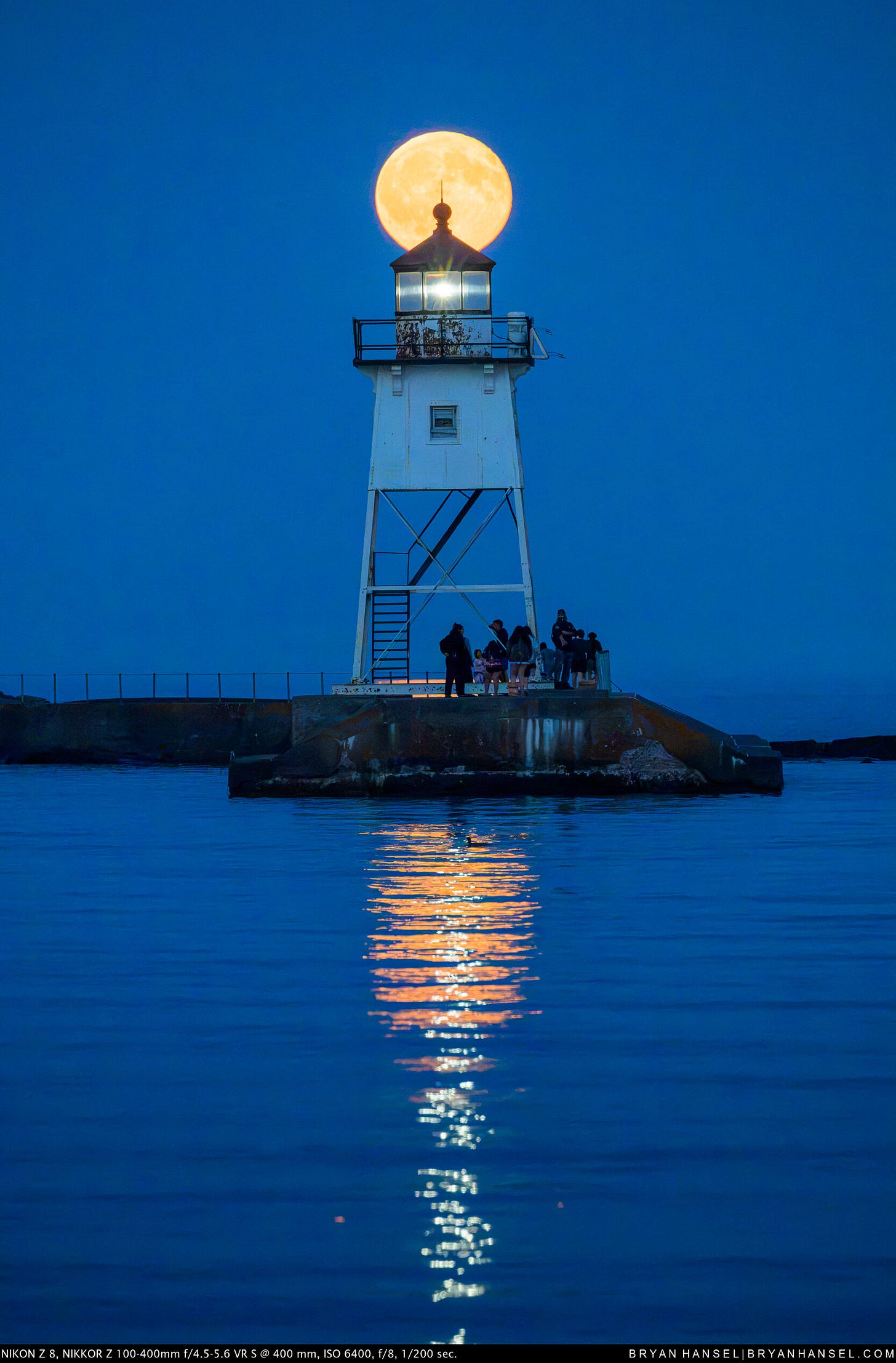A golden moon rises directly behind the lantern room of the Grand Marais lighthouse, casting a bright reflection across the darkening harbor.