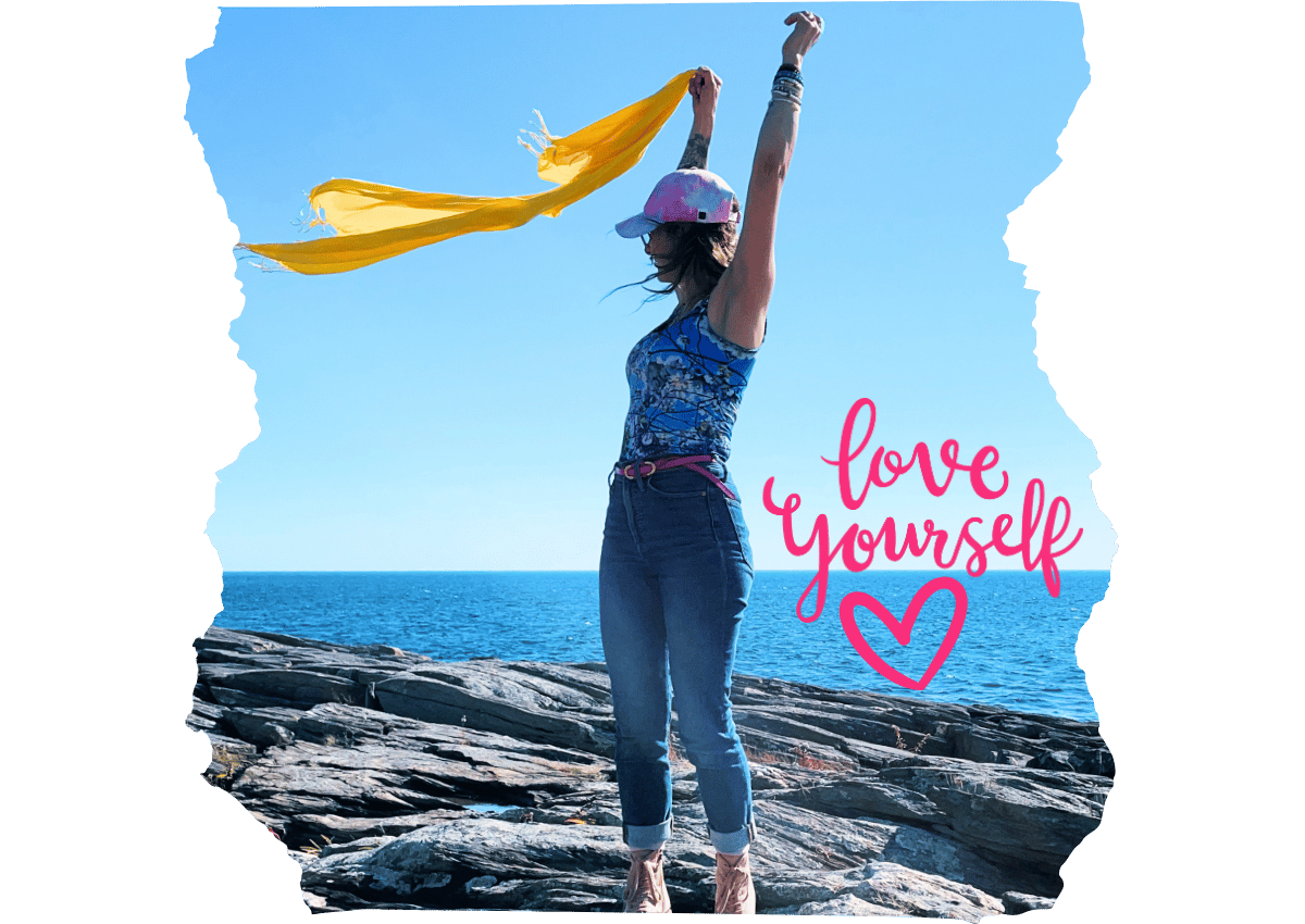 A woman standing on rocky coastal terrain with arms raised, holding a yellow scarf. The image includes the motivational phrase 'Love Yourself' in pink script with a heart, emphasizing self-affirmation."