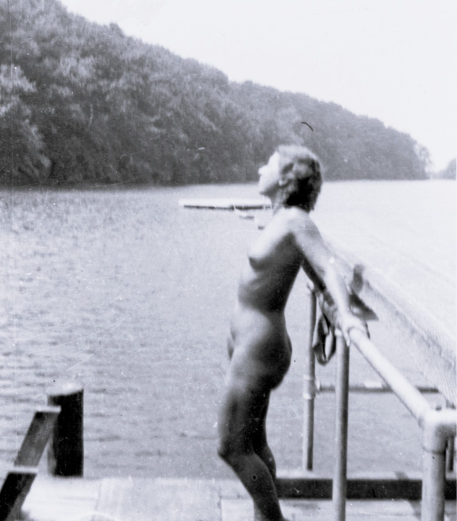 A nude woman stands on a dock beside a lake, leaning back against a railing and looking upward.