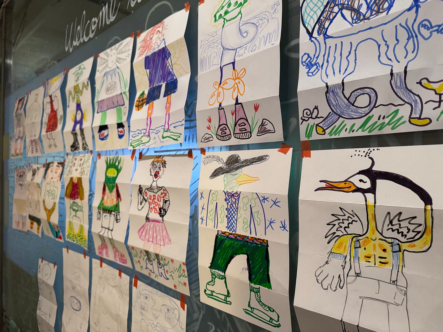 Horizontal photo of many Cadavre exquis works of collaborative surrealist art taped up on a frosted glass wall