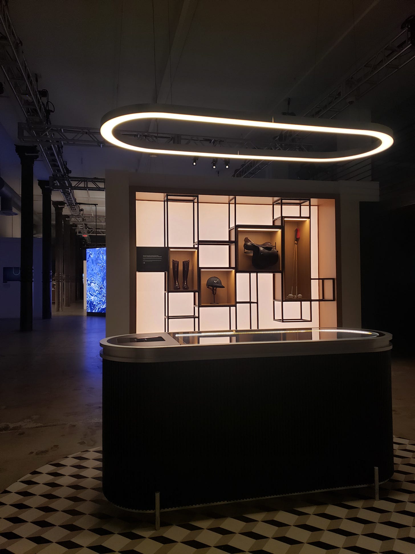Modern exhibition display with a glowing oval ceiling light above a sleek black reception counter. Behind it, a geometric backlit wall showcases polo equipment, including boots, a helmet, a saddle, and a mallet, arranged in open rectangular frames.