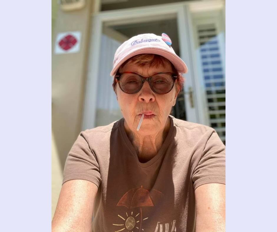 Light purple square background with a picture of a woman, sitting, with a lollipop in her mouth and a pink hat and sunglasses on.