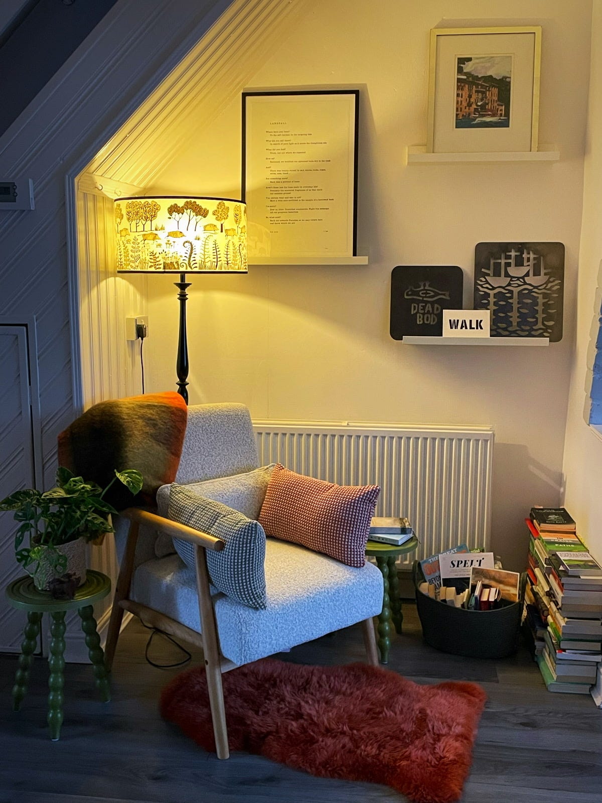 A cozy reading nook with a cream armchair, an orange rug and a small green side table with a leafy houseplant on top. A floor lamp with a cream shade printed with woodland animals casts a warm glow. On the wall behind the chair there’s framed artwork and a poem and a basket next to the chair is overflowing with books and magazines.
