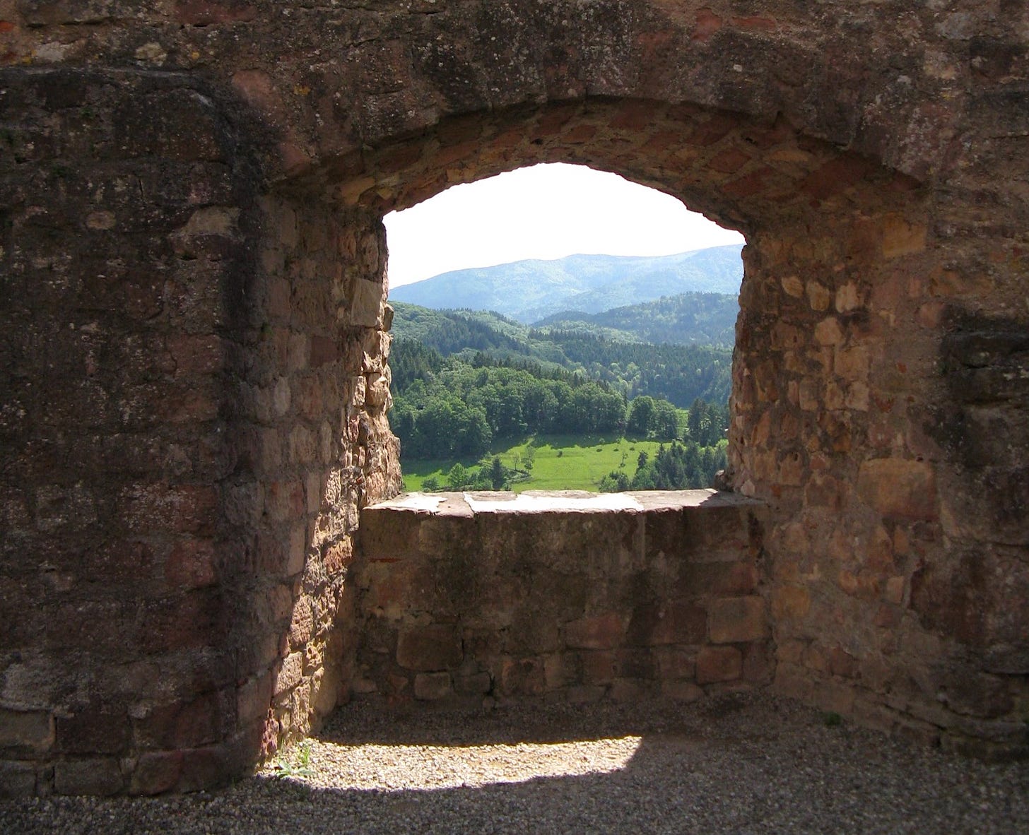 Heavy stonework walls surround an arched stone opening with view of lush green springtime fields, hills, trees