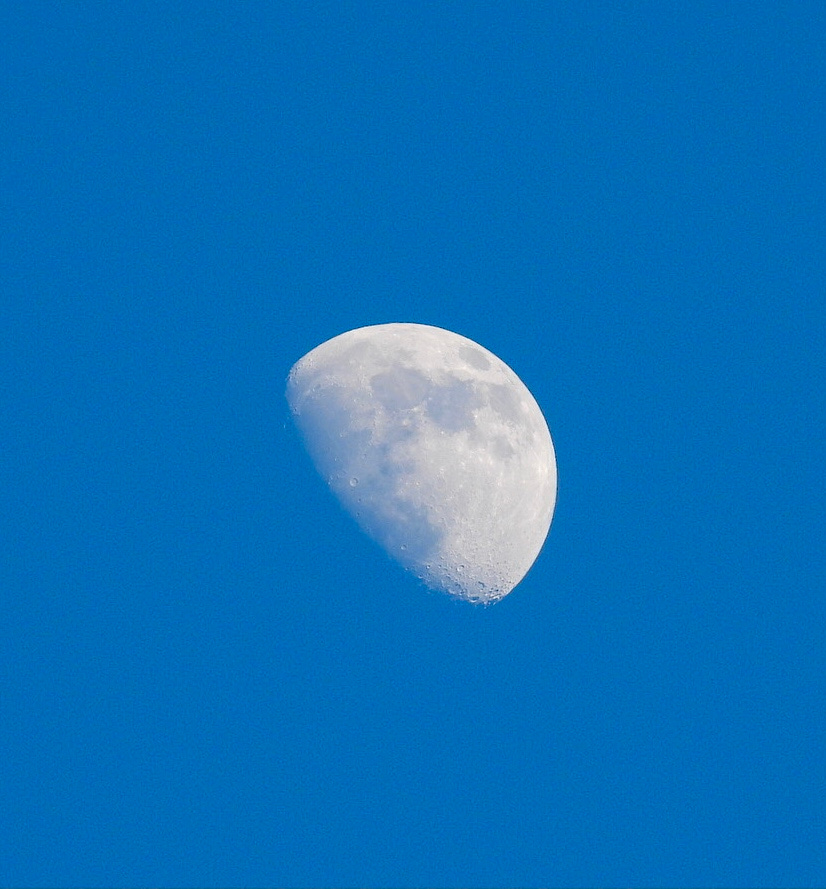 A bright, gray-and-white moon hangs aloft in a deep blue sky. Zoomed in close, the camera captures small craters along the base of the just-past-half-moon, craters that are typically hidden from the naked eye.