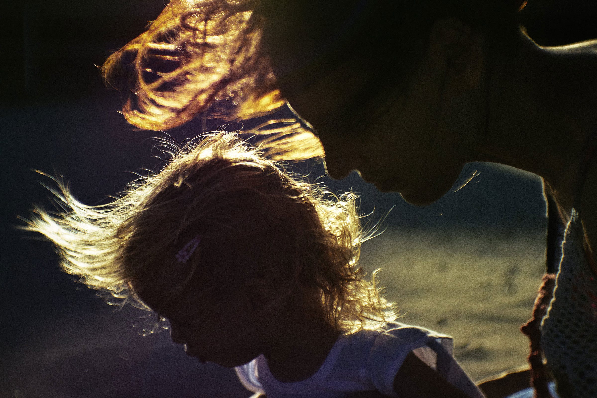 A woman and a young child stand close together in the sunlight, their hair illuminated and blowing in the wind, creating a glowing effect against a dark background.