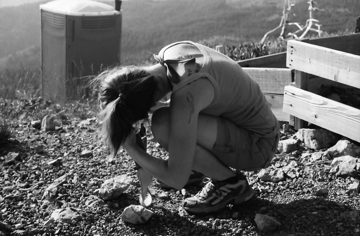 A black and white photo of Sarah, turned away from the camera and curled over in a ball, taking a photo. She is standing on rocky ground and, in the distance, a port-a-potty can be seen.