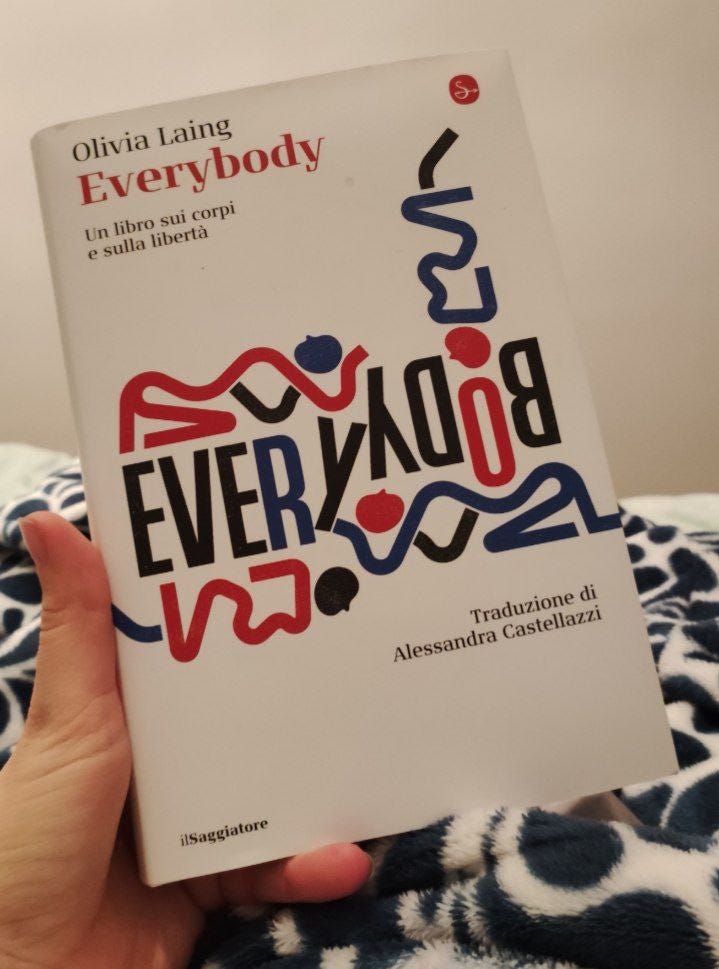 Una mano tiene un libro. Si tratta di Everybody di Olivia Laing. Un disegno compone le lettere del titolo. I colori del libro sono sfondo bianco e scritte in rosso, blu e nero. 
