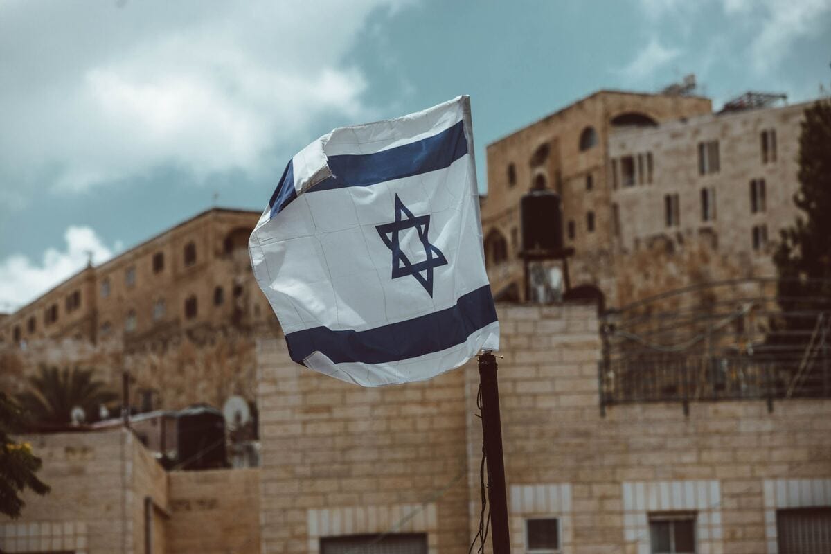 The Israeli flag waves on a sunny day in front of sandy-colored, brick buildings