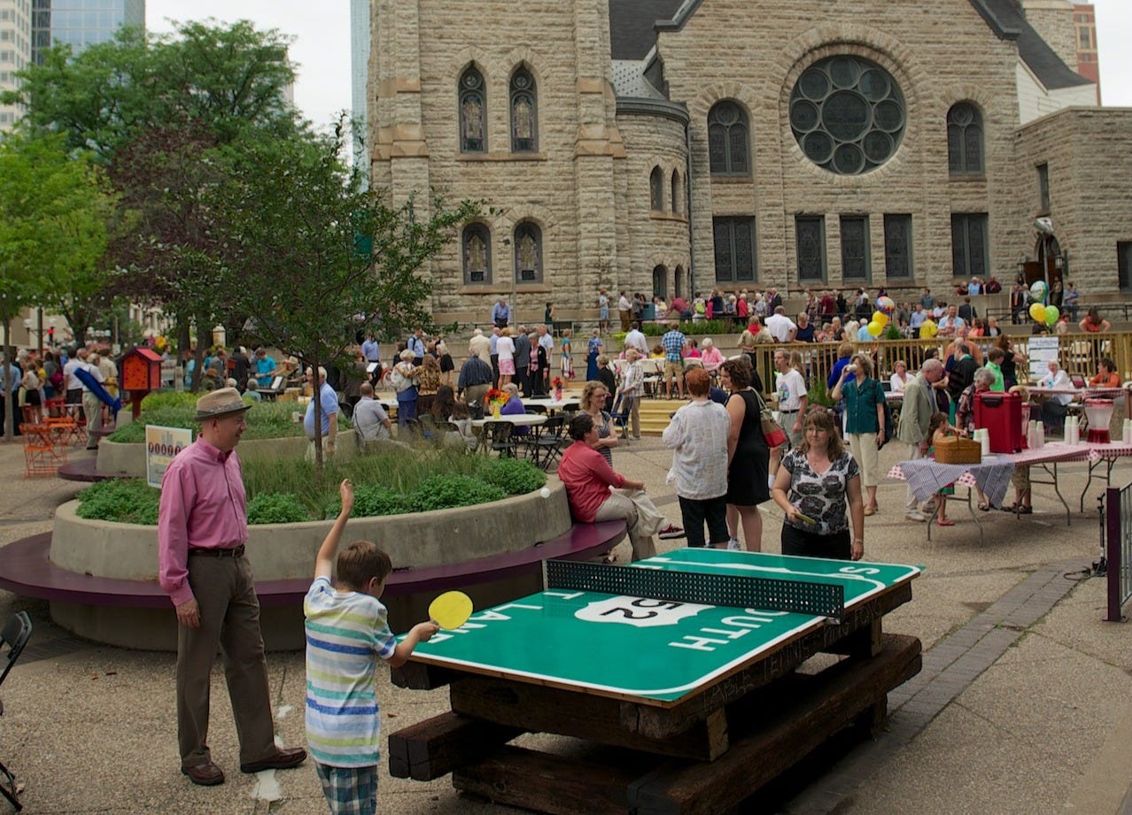 People play ping-pong on a table made from a recycled highway sign. Behind them, people socialize on a wooden deck outside of a cathedral.