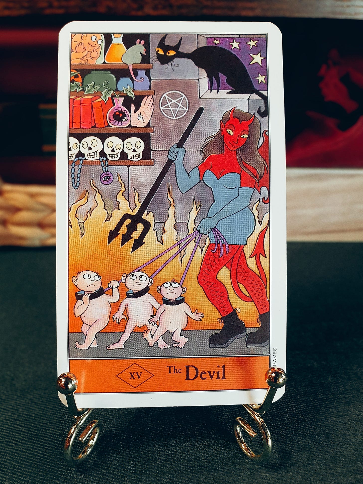 The Devil: The Devil tarot card from Kipling West’s Halloween Tarot, showing a red horned devil holding three small figures on leads amid flames, with skulls, books, a black cat and occult objects in the background. The Devil: The Devil tarot card from Kipling West’s Halloween Tarot, showing a red horned devil holding three small figures on leads amid flames, with skulls, books, a black cat and occult objects in the background.