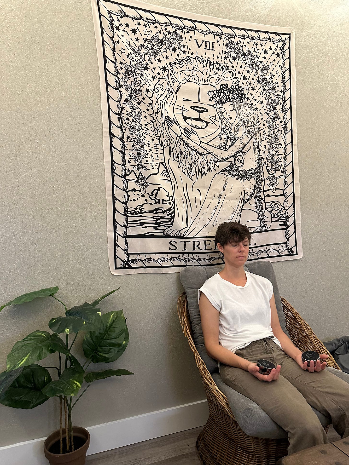 Woman with short brown hair sits in a chair with her eyes shut, holding a black "sound puck" in each hand. There is a large tarot tapestry behind her. Woman with short brown hair sits in a chair with her eyes shut, holding a black "sound puck" in each hand. There is a large tarot tapestry behind her.