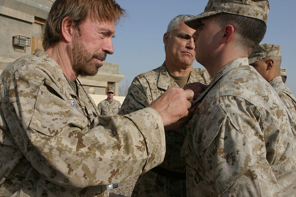 A group of soldiers in desert camouflage uniforms stand outdoors near a concrete building. In the foreground, an older man with a beard carefully pins or adjusts a medal or insignia on a younger soldier’s chest while another senior soldier looks on. Additional personnel stand in the background, and the scene appears to take place in a military setting under clear daylight. A group of soldiers in desert camouflage uniforms stand outdoors near a concrete building. In the foreground, an older man with a beard carefully pins or adjusts a medal or insignia on a younger soldier’s chest while another senior soldier looks on. Additional personnel stand in the background, and the scene appears to take place in a military setting under clear daylight.
