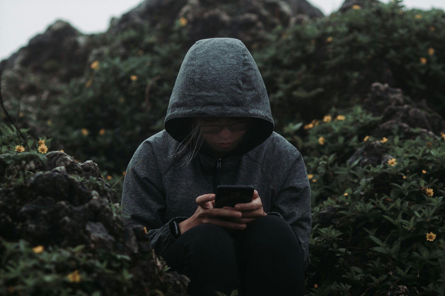 Person in gray hoodie scrolling on smartphone outdoors surrounded by greenery and flowers - social media addiction concept