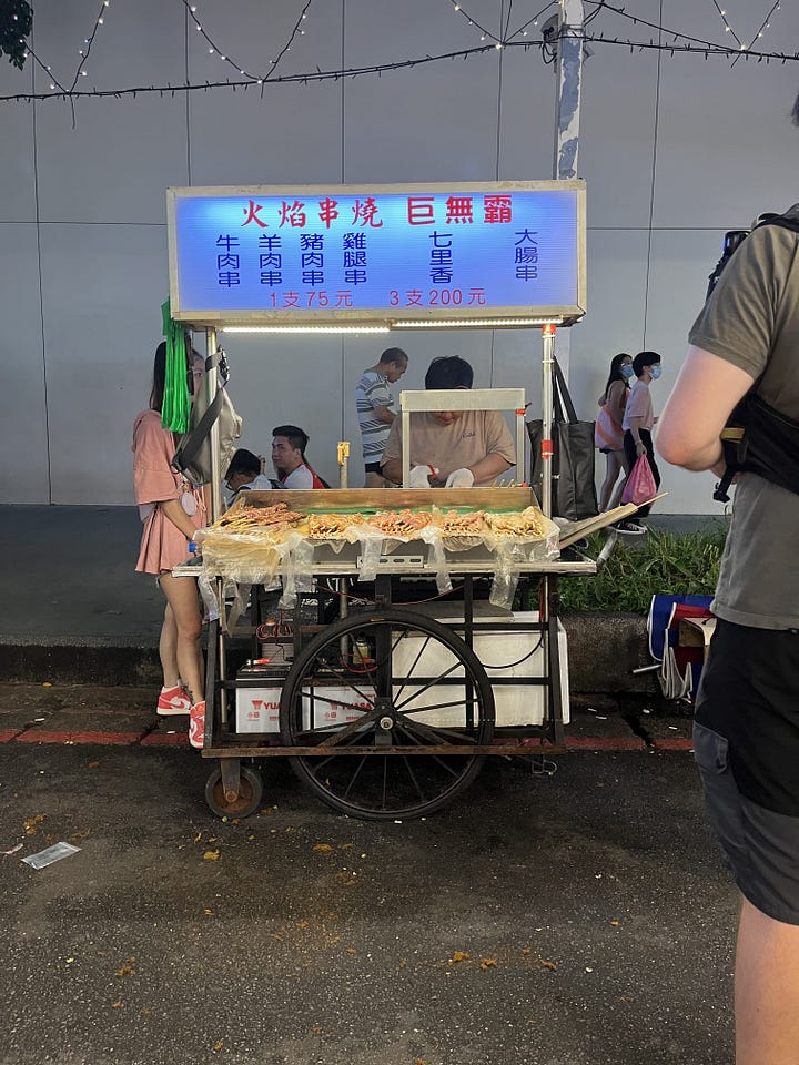 on the left, street stall selling meat skewers. on the right, street stall selling chinese rice cakes
