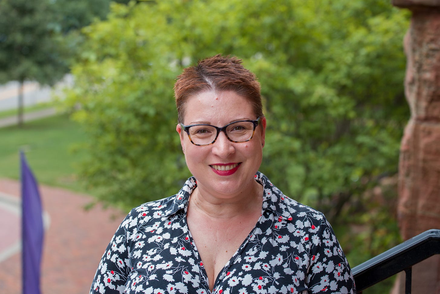 A photo of a white woman with short, auburn hair, smiling at the camera. She wears tortoiseshell glasses, and a black dress with white flowers on it.