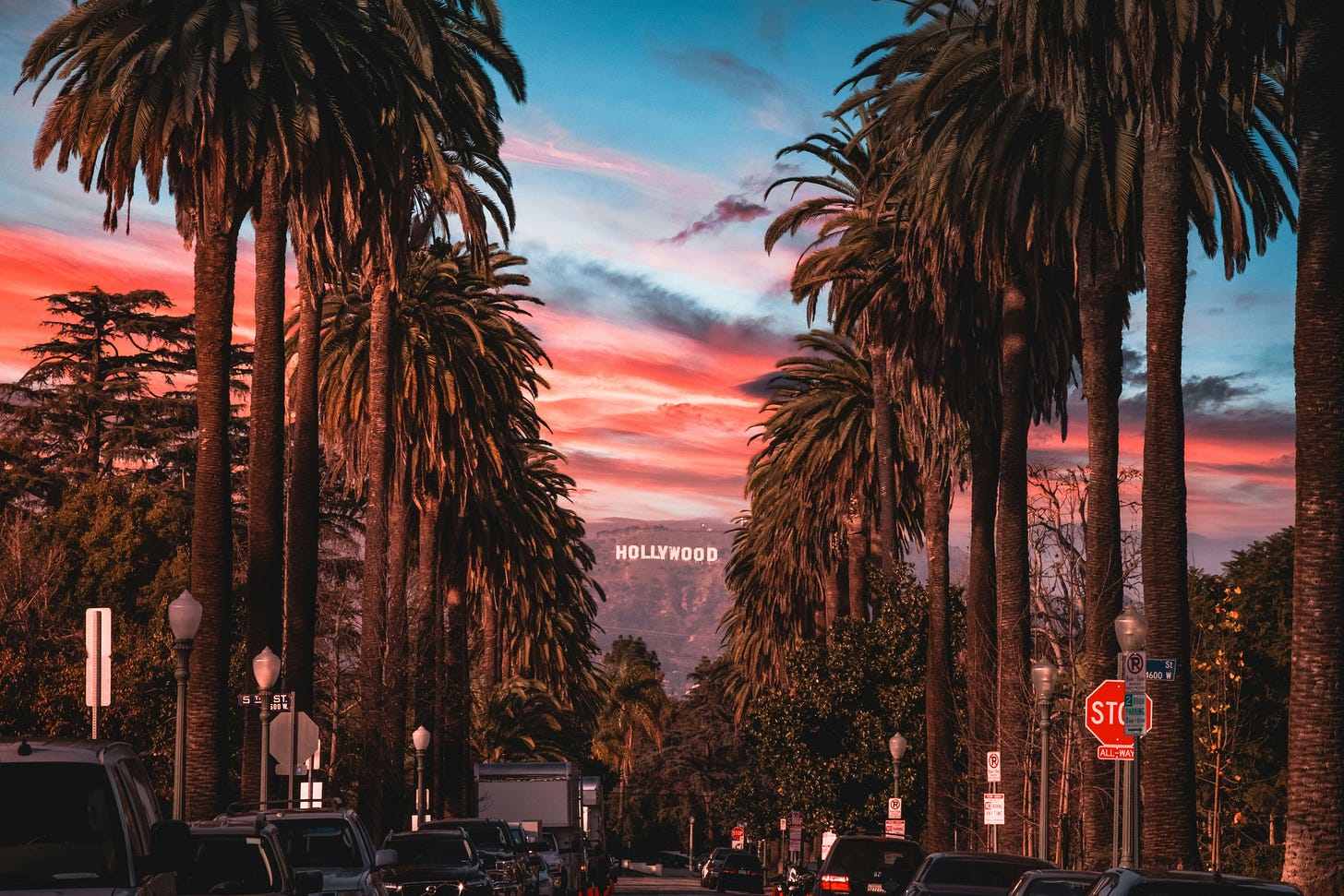 A photograph looking north along a street in Los Angeles lined with tall shaggy palm trees, nearing sunset. The Hollywood sign is visible on a mountainside in the distance, and streaks of orange and black clouds cross a blue sky above.