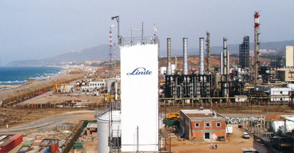A photograph with a large industrial helium processing plant, with multiple smoke stacks and unpaved road crossing into the complex. In the foreground, a tall white building bears the logo for Linde, the German multinational oil and gas company. On the left of the photo, behind the plant there is a coastline of an ocean or sea, and on the horizon behind the plant, with out of focus in the far distance, some hills and possibly other buildings or a town.