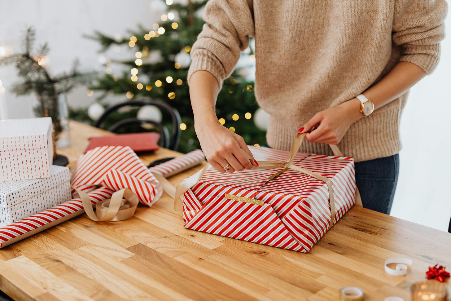 “A woman wrapping striped Christmas gifts on a wooden table with a tree in the background, reflecting the time and care that goes into holiday giving.”