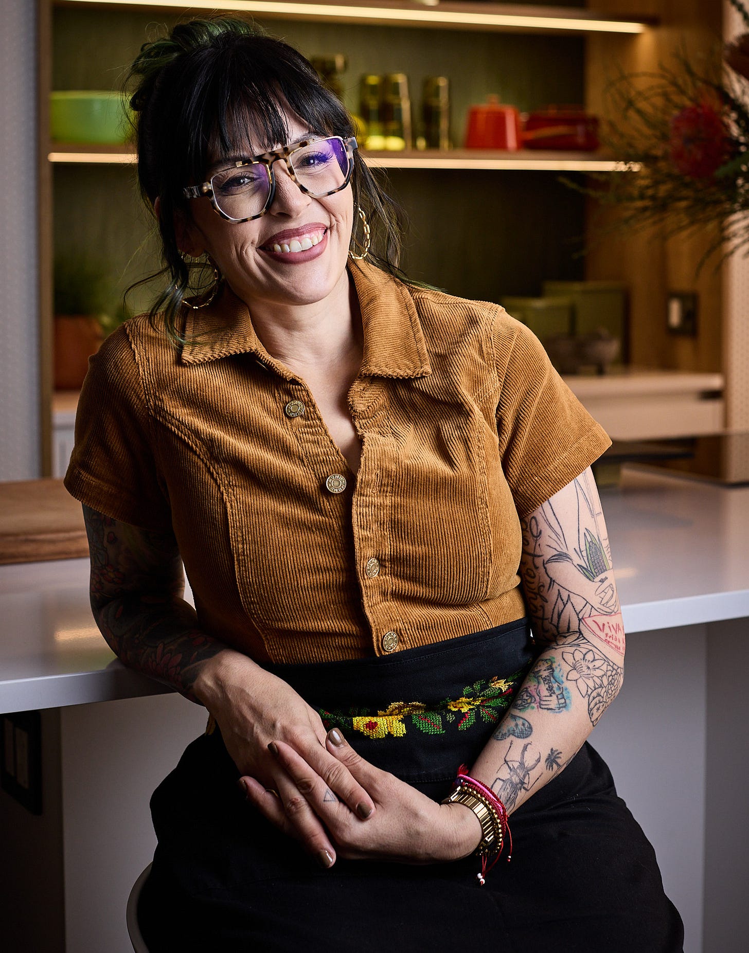 Claudette Zepeda, a woman with dark brown hair put up in a bun, tortoiseshell glasses, and dark red lipstick, leaning against a countertop
