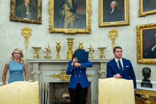 Gov. Gretchen Whitmer of Michigan holding blue binders in front of her face as she stands in the Oval Office, with two Trump aides flanking her. In the background are a fireplace, gold-framed paintings of presidents and other gold decorations.