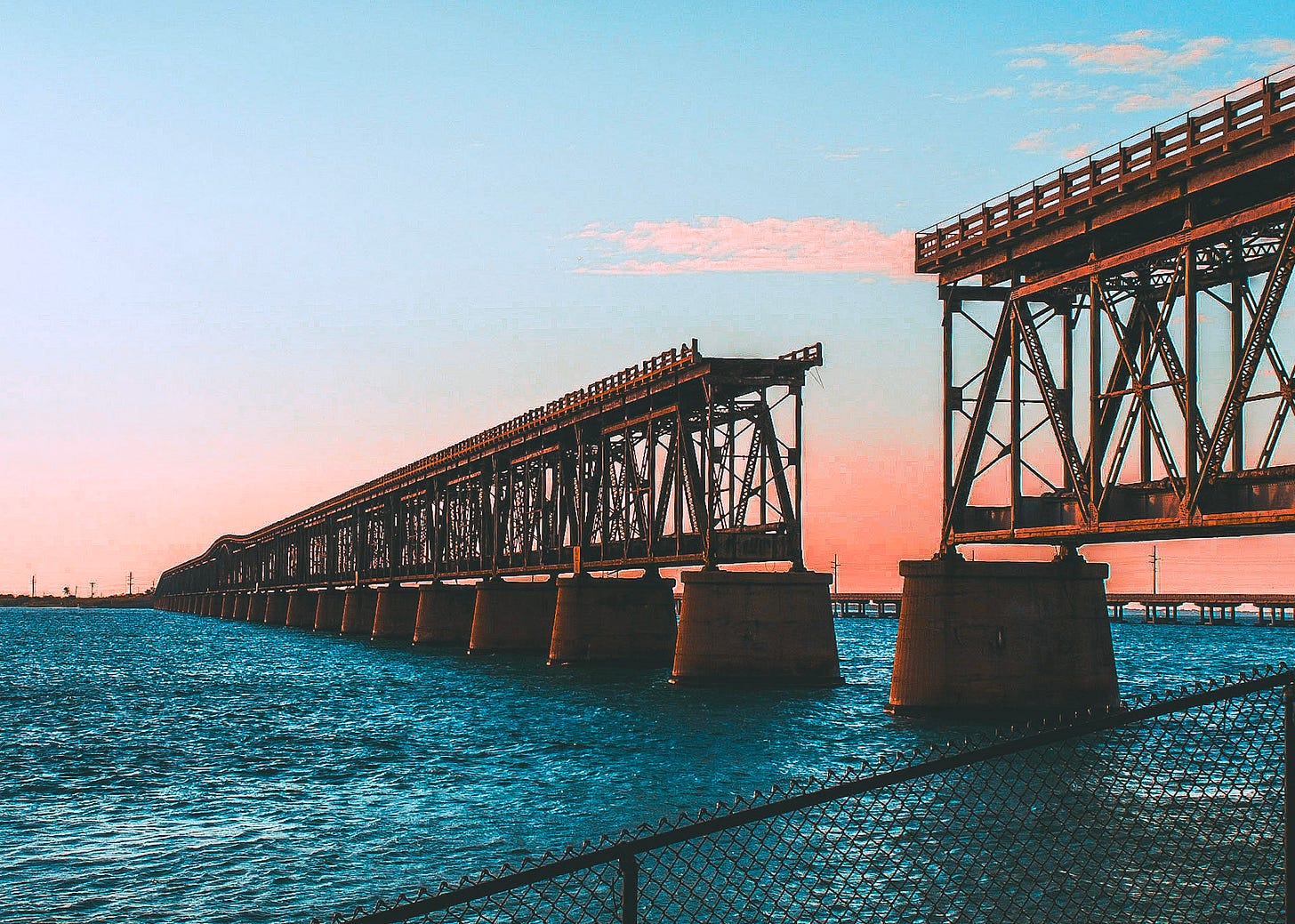 A long, abandoned, broken railway bridge stretches across bright blue ocean water towards a pink and orange sunset sky. The decaying metal structure and concrete supports are visibly corroded and in disrepair, with a section of the bridge missing in the foreground.