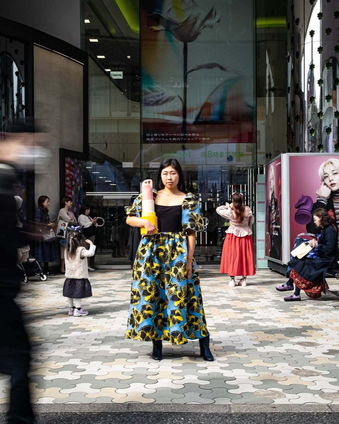 Artist Eri Maeda stands in the center of Laforet Harajuku during Art Week Tokyo 2024, holding a bold ceramic sculpture. She wears a patterned dress, surrounded by passersby in a busy urban scene blending art, fashion, and everyday life. Artist Eri Maeda standing outside Laforet Harajuku holding a ceramic sculpture, surrounded by passersby during Art Week Tokyo 2024. Contemporary art in a busy urban fashion setting.