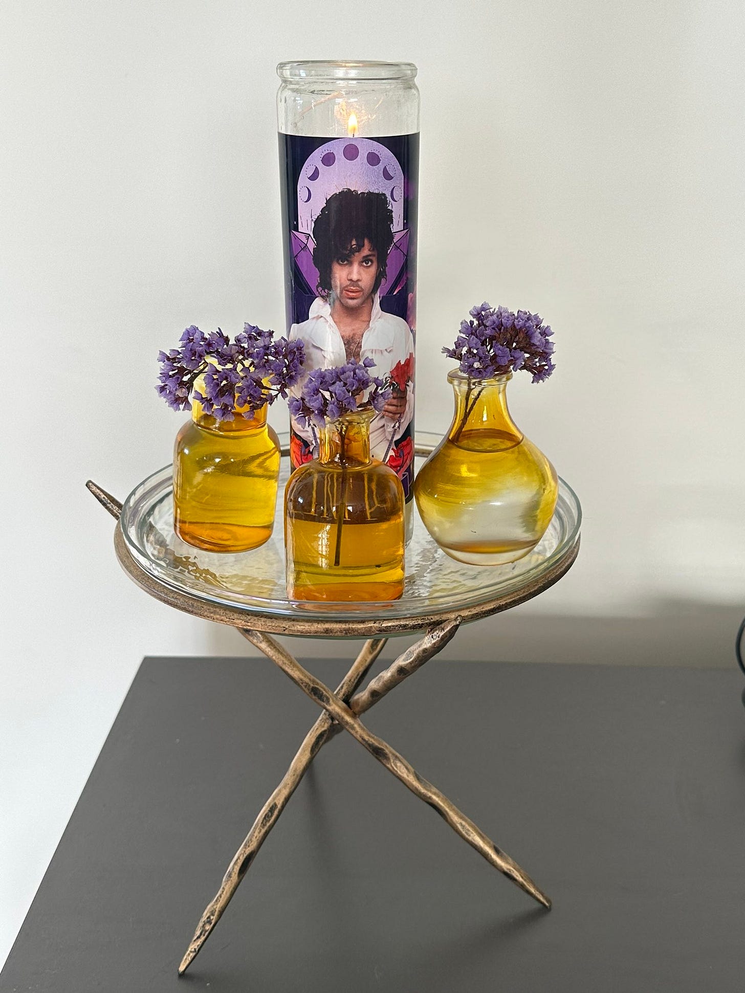 A top a glass plate sits a lit Prince prayer candle. Surrounding it are three small yellow jars with purple flowers in them.