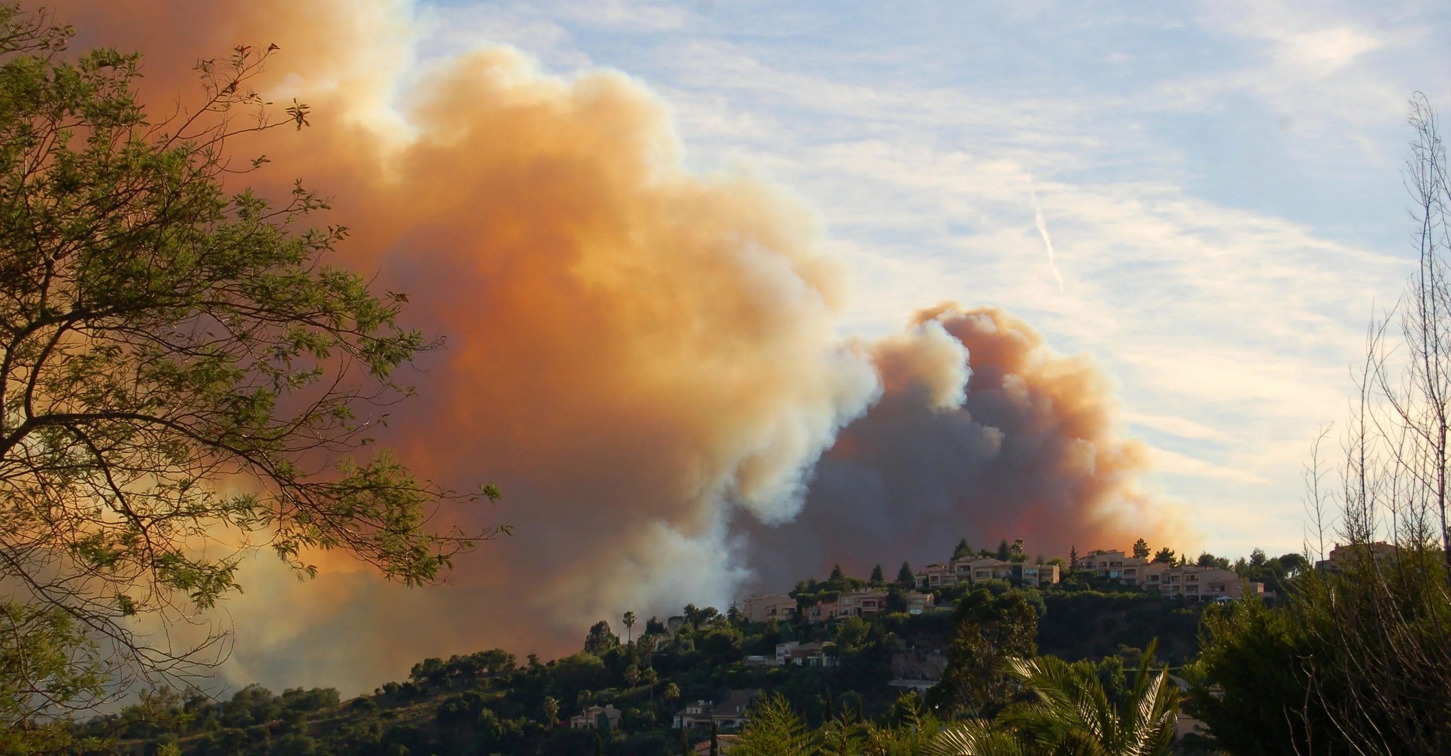 Panoramic view of French hillside, homes endangered. Orange and umber clouds of smoke contrast a blue sky.