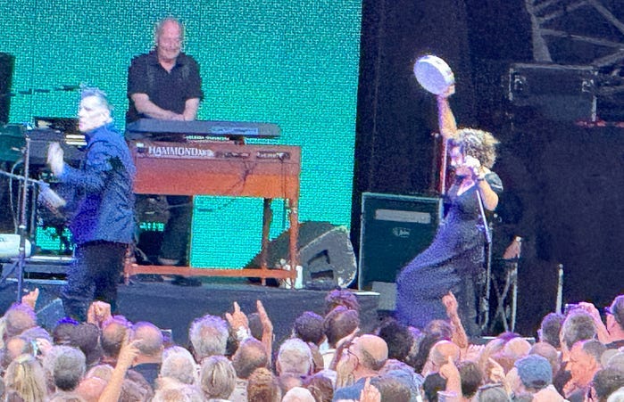 On stage are Deacon Blue 2 males and 1 female who is dancing and has a tamborine raised in the air.