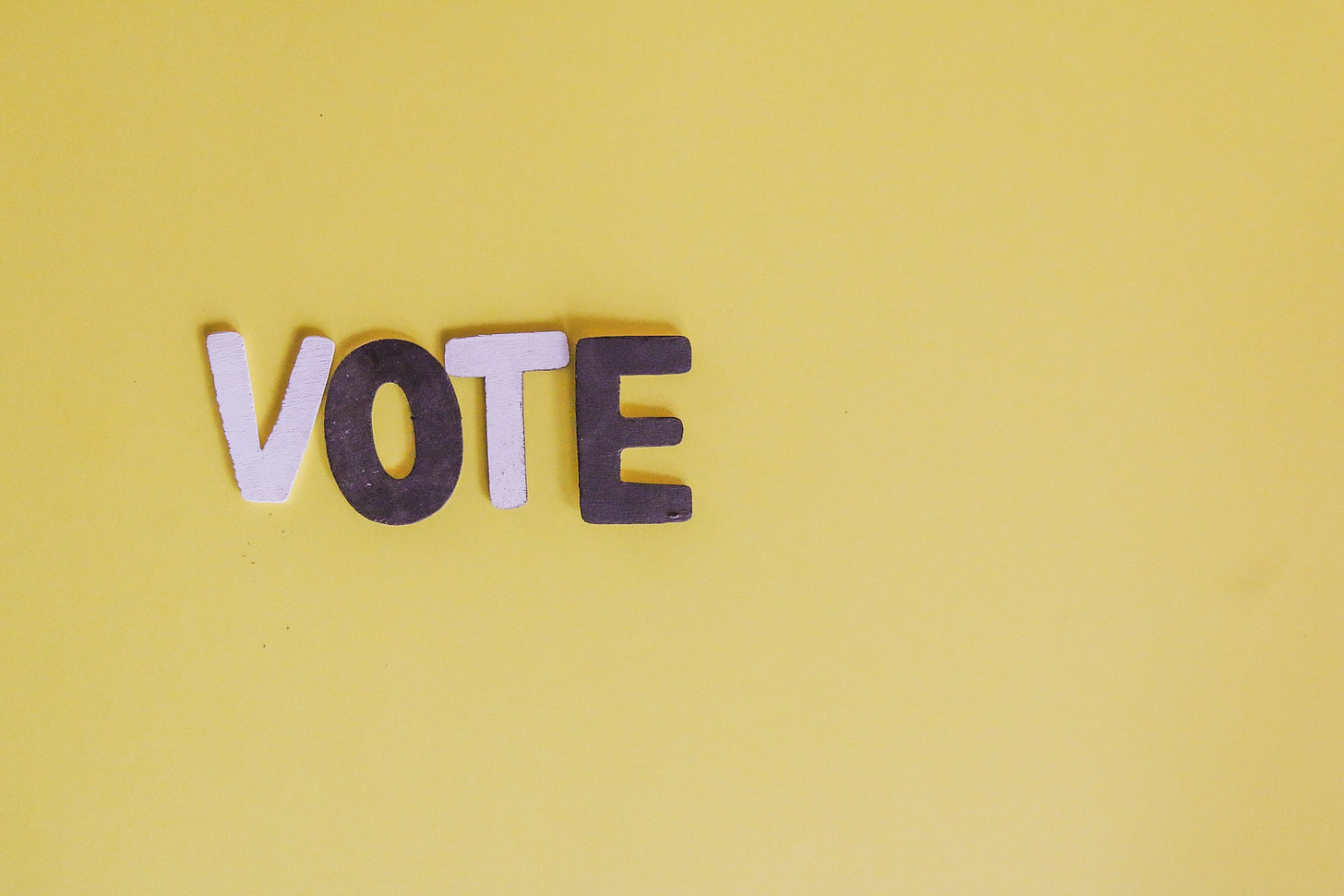 La parola "Vote" con lettere rosa e viola appoggiate su uno sfondo giallo ocra. La parola "Vote" con lettere rosa e viola appoggiate su uno sfondo giallo ocra.