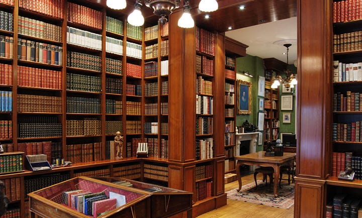 Interior gallery of Peter Harrington Rare Books in London. The first image shows a warm wood-paneled library with floor-to-ceiling mahogany shelves of leather-bound antiquarian books in red, green, and tan calf and morocco bindings, a glass-topped display cabinet of rare first editions, and a green-walled reading room beyond with a marble fireplace, oil portrait, and antique writing table. The second image shows a brighter showroom with tall glass-fronted display cases holding open antiquarian books, hand-colored maps, and illustrated plates above lower shelves of gilt leather-bound sets, with polished hardwood floors running into an adjoining room.