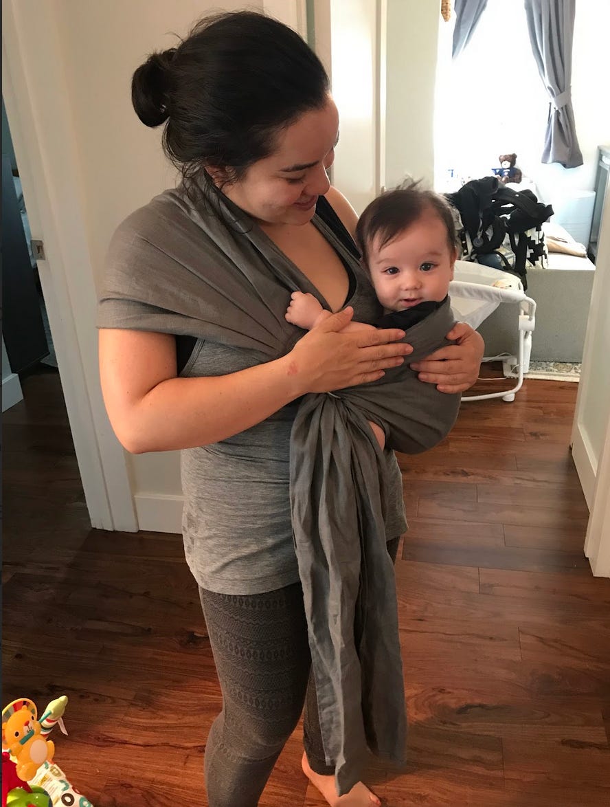 Corinne holding baby; Asian woman in comfortable outfit with baby sling; baby loking at camera; they are in a house, you can see a toy on the floor and Corinne holding baby; Asian woman in comfortable outfit with baby sling; baby loking at camera; they are in a house, you can see a toy on the floor and