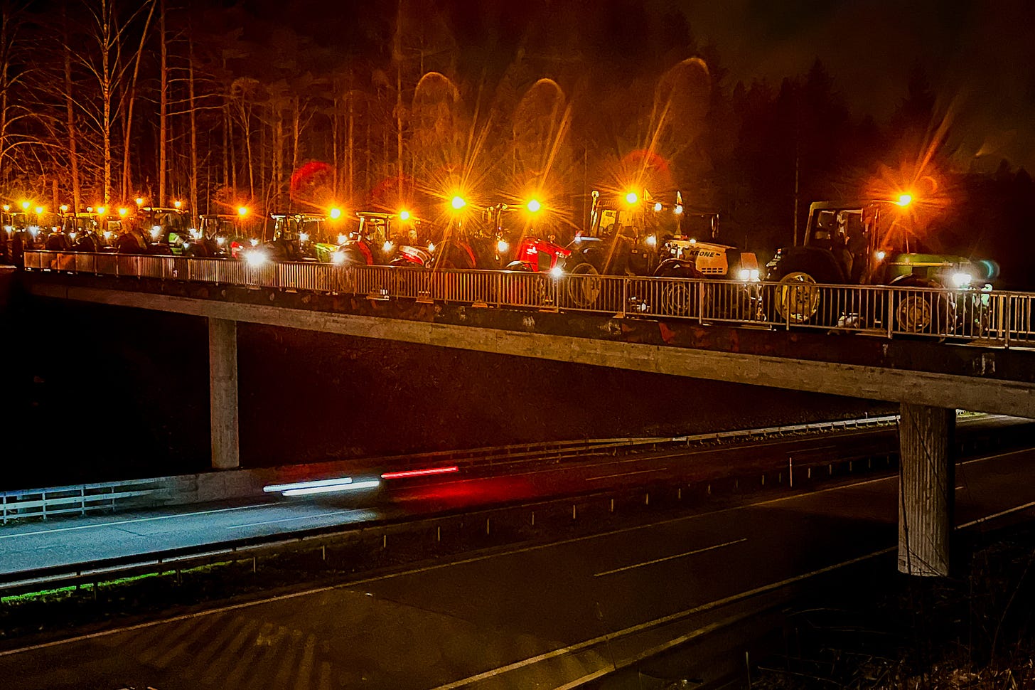 Bei einem nächtlichen Bauernprotest fährt eine lange Kolonne von Traktoren mit eingeschalteten Scheinwerfern und orangefarbenen Rundumleuchten über eine Brücke. Unter der Brücke verläuft eine Autobahn, auf der vorbeifahrende Autos als Lichtspuren zu sehen sind; im Hintergrund zeichnet sich ein dunkler Wald ab.